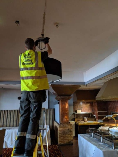 Workman in yellow high-fis is fitting a big light in a kitchen. He is on the left hand side on a ladder.