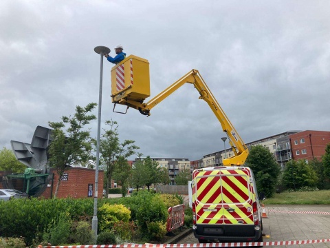 A van with yellow and red arrows on its back has a yellow crane attached, and is holding a man inside who is fixing or attaching a pole, outdoors. The location has plenty of green shrubbery and trees around, and housing and buildings in the background. In the foreground and at the bottom there is some red and white tape going around, to cordon off the area so pedestrians don't walk there while it gets fixed.