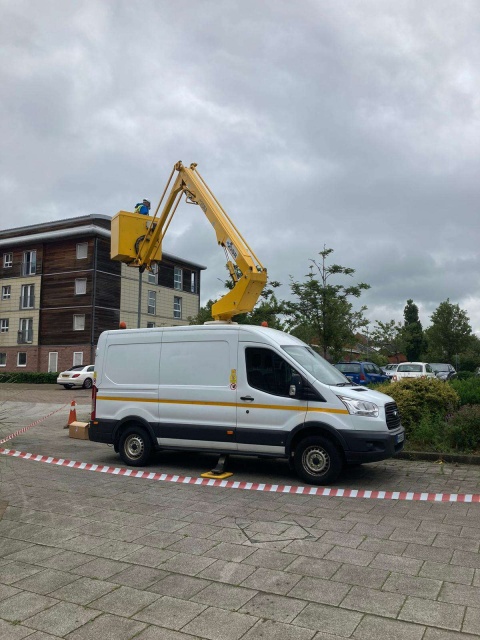 this image is taken outdoors. It is focused on a white van with a yellow crane on it which has a workman inside, going to fix an electrical pole outdoors. It is cordoned off with red and white tape. There is green and yellow shrubbery and trees in the background on the right, and on the left there is a block of houses/offices.