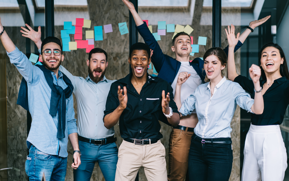 A group of employees all cheer in front of an open office building. They are happy and motivated.